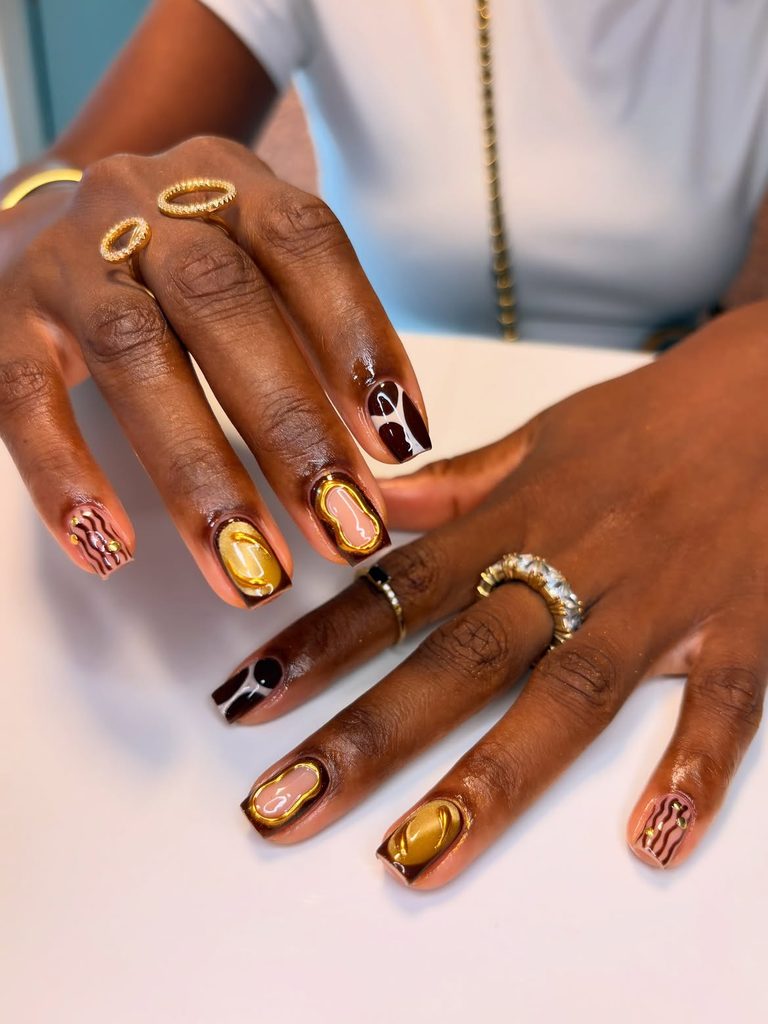 Short square nails with chocolate brown, gold frames, gold shimmer, and wavy line accents on deep skin tone