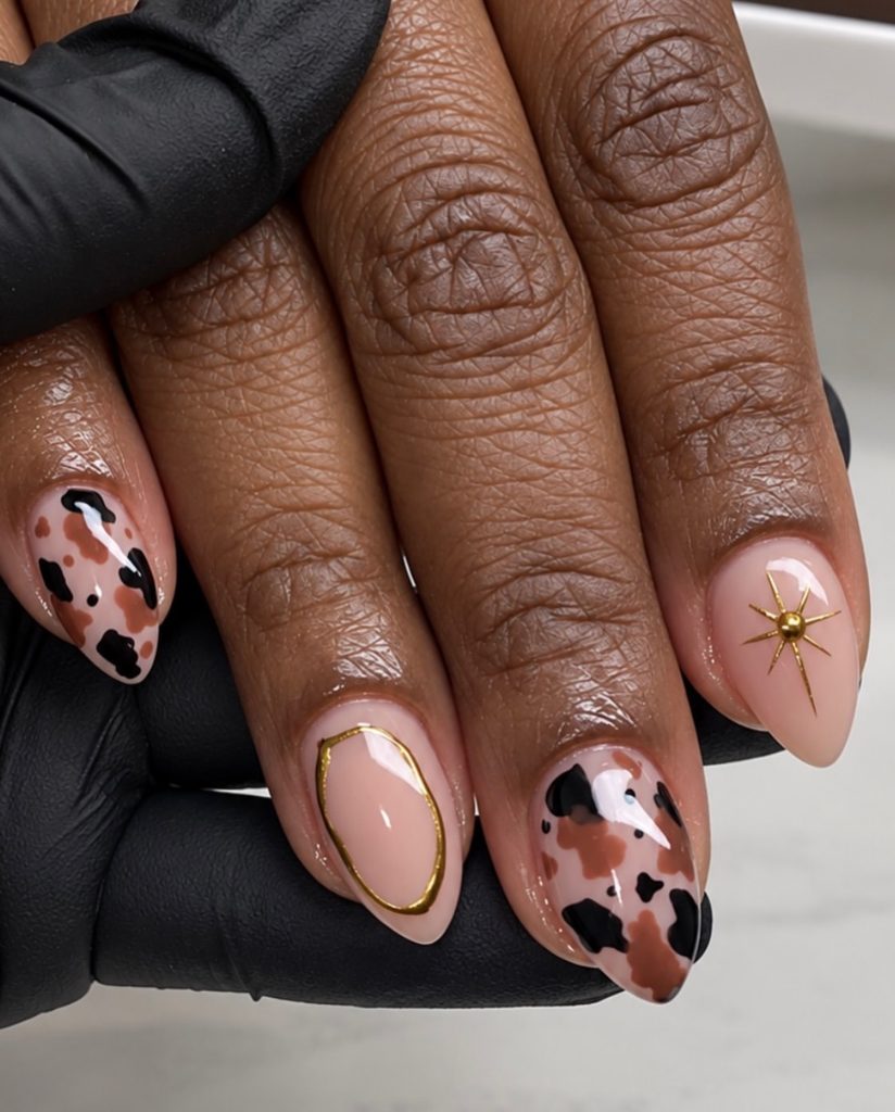 Almond nails with sheer nude base, black and brown cow print, gold oval outline, and gold starburst